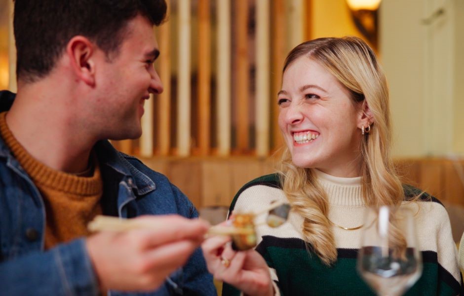 Casal sorridente desfrutando de um momento romântico em restaurante, com destaque para a mulher com cabelo loiro e o homem com cabelo castanho, em um ambiente acolhedor.
