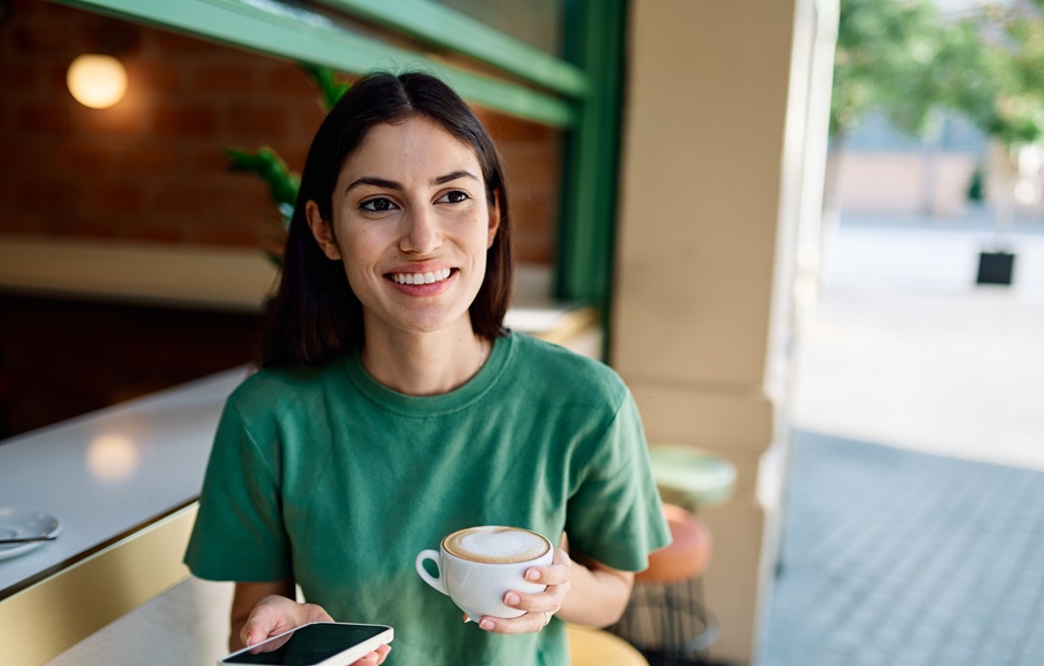 Mulher sorridente com cabelo castanho escuro, vestindo uma camiseta verde, segurando uma xícara de café ou chá, sentado em um café ou restaurante bem iluminado, com ambiente acolhedor e decorado com elementos naturais.