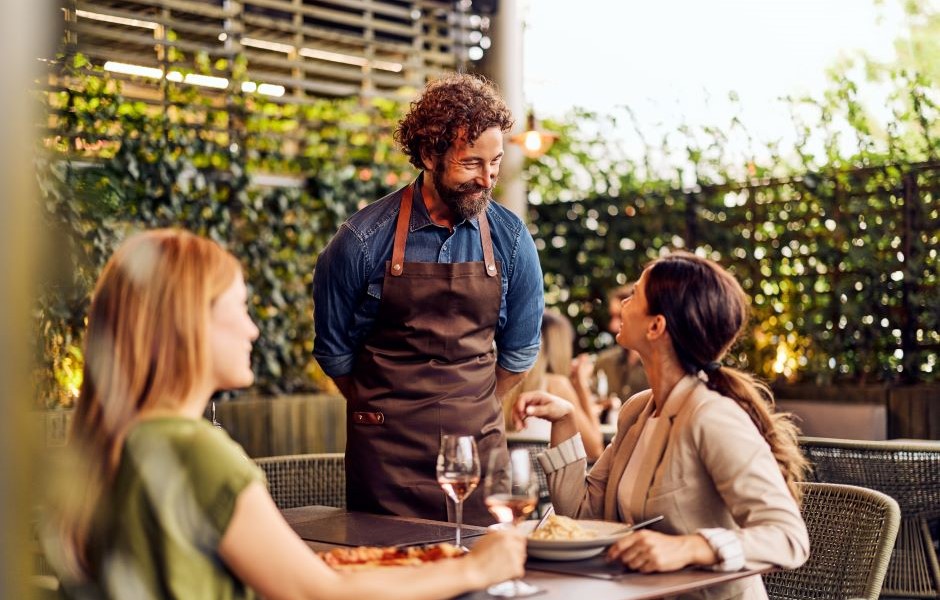 Homem com barba e cabelo cacheado atendendo duas mulheres em um restaurante ao ar livre, com pratos de comida e taças de vinho, ambiente acolhedor e natural.