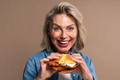 Mulher idosa feliz e sorridente segurando um hambúrguer de frango, destacando a alegria de comer fast food em um fundo neutro.