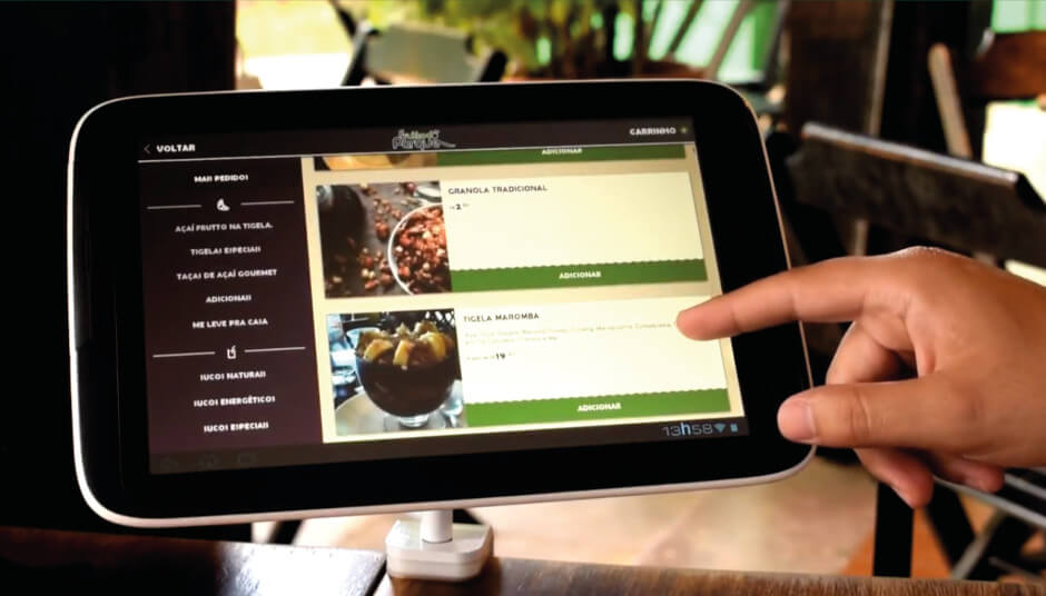 Imagem de um tablet exibindo um cardápio digital com opções de comida, incluindo granola tradicional e tigela de açaí, em um ambiente aconchegante.