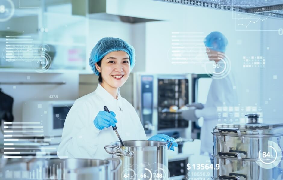 Imagem de uma cozinheira em uma cozinha industrial moderna, com utensílios de cozinha e tecnologia visível.