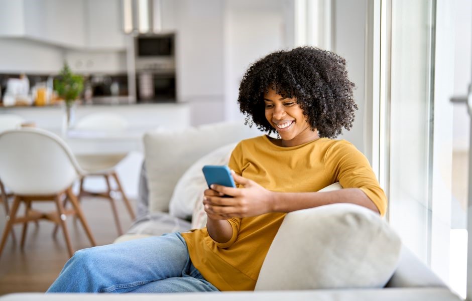 Mulher sorrindo usando celular no sofá em casa, ambiente moderno e aconchegante com iluminação natural, ideal para postar sobre comunicação e tecnologia.