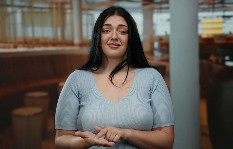 Mulher com cabelo preto e expressão de dúvida ou insegurança, vestindo camiseta azul clara, posando em ambiente de restaurante ou cafeteria.