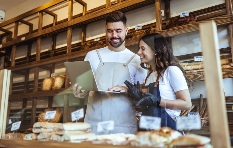Duas pessoas no balcão de uma padaria, um homem e uma mulher, sorrindo e observando uma tabela de produtos, com prateleiras de pães ao fundo, demonstrando uma conversa descontraída.