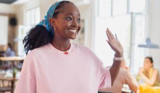 Jovem mulher africana sorridente com cabelo cacheado, usando uma blusa rosa e uma headband azul, em ambiente social, gesticulando com a mão ao lado, transmitindo alegria e entusiasmo.