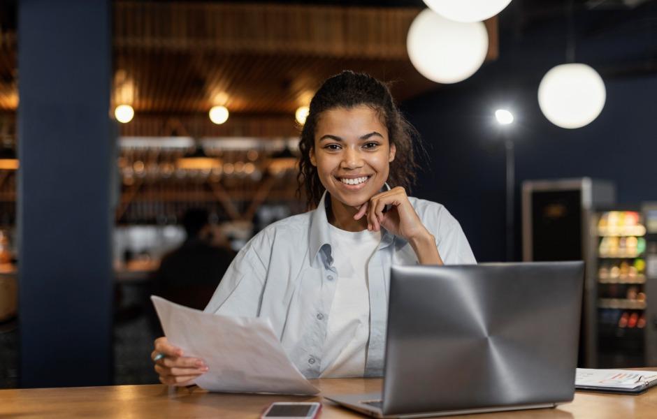 Mulher sorridente sentada à mesa com notebook e segurando papéis em um ambiente de escritório ou café.