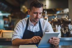 Homem de avental concentrado usando um tablet sobre um balcão de atendimento. O fundo mostra a cozinha de um restaurante ou café.