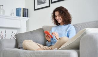 Jovem sorridente usando celular enquanto relaxa no sofá de casa, ambiente moderno e aconchegante, ideal para o conforto e entretenimento.