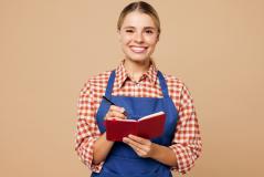 Mulher sorridente usando avental azul segurando um caderno vermelho e uma caneta, em um fundo neutro, representando o dia a dia de um trabalhador. Ideal para conteúdos sobre trabalho e organização.