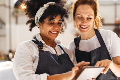 Duas mulheres sorrindo e olhando para um tablet em uma cozinha moderna, vestindo aventais pretos, o que indica uma aula de culinária ou cozinha colaborativa.