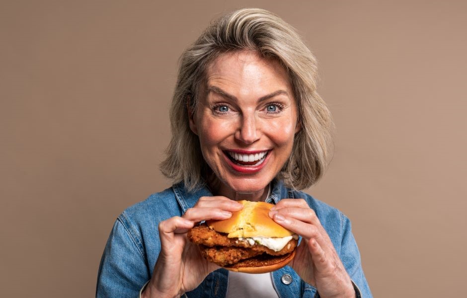 Mulher idosa feliz e sorridente segurando um hambúrguer de frango, destacando a alegria de comer fast food em um fundo neutro.