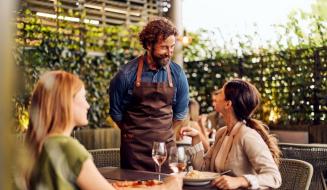 Homem com barba e cabelo cacheado atendendo duas mulheres em um restaurante ao ar livre, com pratos de comida e taças de vinho, ambiente acolhedor e natural.