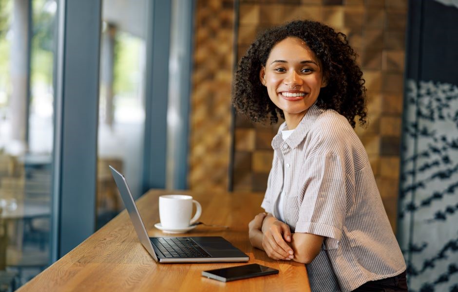 Mulher sorridente com cabelo cacheado no ambiente de um escritório, usando camisa listrada, ao lado de um laptop, uma xícara de café e um celular, representando profissionalismo e produtividade.