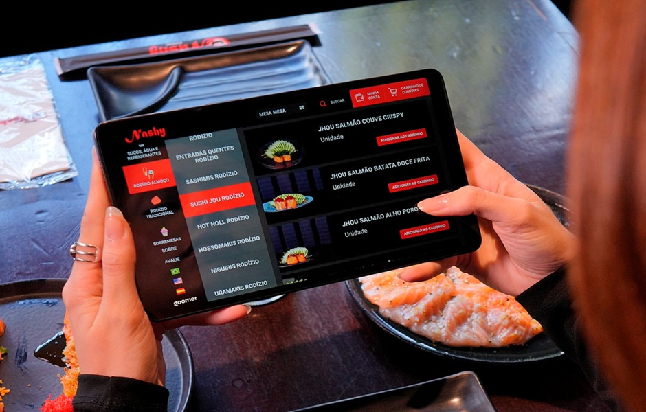 Pessoa usando um tablet para fazer pedido em um restaurante de comida japonesa, com pratos de sashimi e salmão visíveis na mesa, destacando o uso de tecnologia na experiência gastronômica.