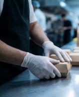 Profissional de luvas organiza embalagens de papel kraft em uma linha de montagem de delivery dentro de uma cozinha industrial movimentada.