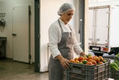 Mulher com avental e touca segura uma caixa plástica com tomates e pimentões em um entreposto ou cozinha comercial, com um homem e um caminhão de entregas ao fundo.