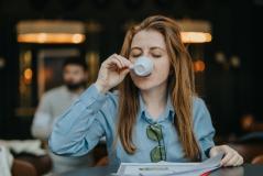 Mulher com cabelo castanho, usando blusa azul e óculos de grau presos à camisa, tomando uma xícara de café ou chá em um ambiente aconchegante, com foco na expressão de satisfação. Ao fundo, pessoas estão em um espaço moderno e bem iluminado, criando uma atmosfera de cafeteria ou sala de reunião, ideal para quem busca conforto e relaxamento.