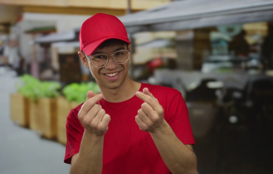 Jovem sorridente usando boné vermelho e óculos transparentes faz gesto com as mãos enquanto está em um ambiente ao ar livre, promovendo estilo casual.