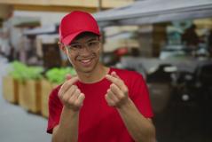 Jovem sorridente usando boné vermelho e óculos transparentes faz gesto com as mãos enquanto está em um ambiente ao ar livre, promovendo estilo casual.