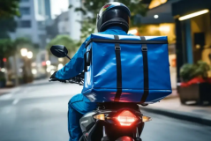 Entregador em moto com baú azul circulando por rua da cidade.