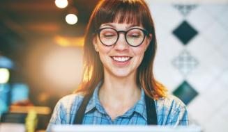 Mulher sorridente com óculos de armações pretas e cabelo castanho, usando camisa xadrez azul, em ambiente de cafeteria, transmitindo alegria e satisfação.
