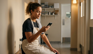 Pessoa de cabelo encaracolado apanhado, t-shirt escura e avental bege utiliza telemóvel sentada em banco de madeira em área de descanso ou corredor de estabelecimento comercial. Ao seu lado encontra-se garrafa de água, enquanto ao fundo observam-se prateleiras de arrumação, portas e iluminação ambiente suave.