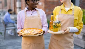 Duas mulheres sorridentes usando aventais, uma segurando uma pizza e a outra uma jarra de limonada, em ambiente ao ar livre, promovendo comida caseira.