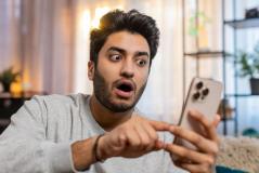 Homem surpreso usando celular em casa, ilustrando a experiência de diversão e surpresa ao usar um smartphone com tecnologia moderna.