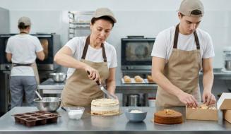 Três confeiteiros de camiseta branca, avental bege e boné trabalham em uma cozinha industrial. No primeiro plano, uma mulher espalha recheio em um bolo com espátula enquanto um rapaz embala doces em uma caixa de papelão. Ao fundo, uma terceira pessoa trabalha de costas perto de fornos e prateleiras de inox.
