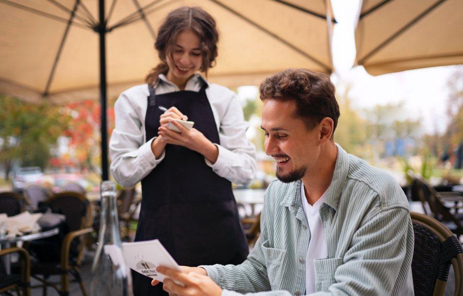 Garçonete anotando o pedido de um cliente sorridente que analisa o cardápio em um restaurante ao ar livre.