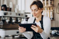 Barista sorridente de avental preto opera um tablet com as duas mãos em um balcão de cafeteria. Ao fundo, destaca-se uma máquina de café expresso profissional branca.