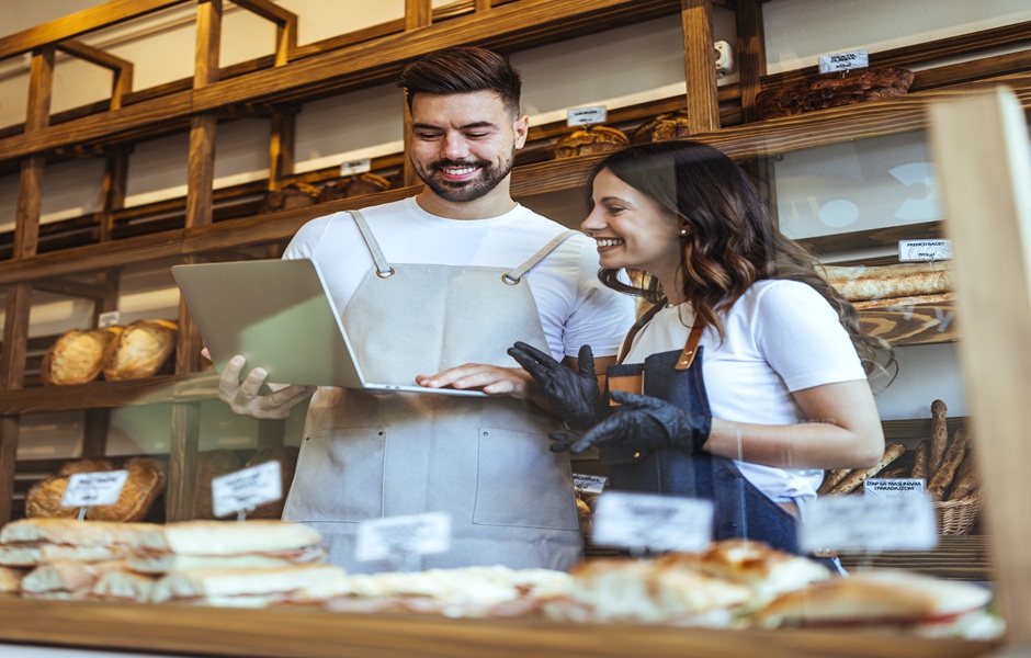 Duas pessoas no balcão de uma padaria, um homem e uma mulher, sorrindo e observando uma tabela de produtos, com prateleiras de pães ao fundo, demonstrando uma conversa descontraída.