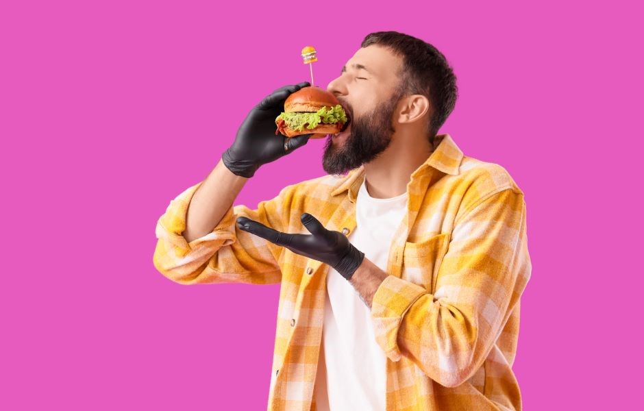 Homem jovem com barba e cabelo curto, usando jaqueta xadrez amarela e branco, segurando um hambúrguer com uma luva preta, pronto para comer, contra um fundo rosa vibrante.