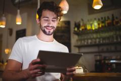 Homem sorridente em uma cafeteria ou bar, segurando e olhando para um cardápio digital em um tablet.