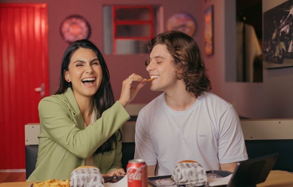 Casal sorridente compartilhando um momento divertido em uma lanchonete, saboreando comida e bebida, com um ambiente descontraído ao fundo.