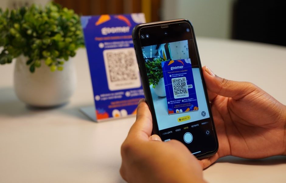 Pessoa escaneando um código QR com um celular na mesa, com uma embalagem azul ao fundo, promovendo a leitura de QR code para acesso a informações e ofertas.