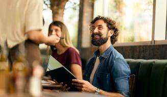Homem sorridente em um restaurante ou café, interagindo com um garçom enquanto lê um cardápio, ao lado de uma mulher. Ambiente aconchegante com grande janela.