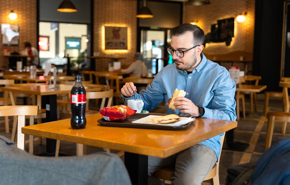 Homem com barba e óculos tomando café da manhã com pão e refrigerante em cafeteria moderna e aconchegante.