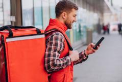 Entregador de colete vermelho e camisa xadrez, visto de perfil em uma calçada, carrega uma grande mochila de delivery nas costas enquanto mexe em seu smartphone.