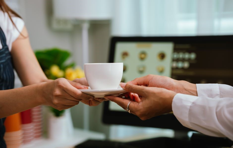 A mulher recebendo uma xícara de café de um atendente em um ambiente interno, com computador ao fundo, simbolizando serviço de cafeteria ou entrega de bebidas