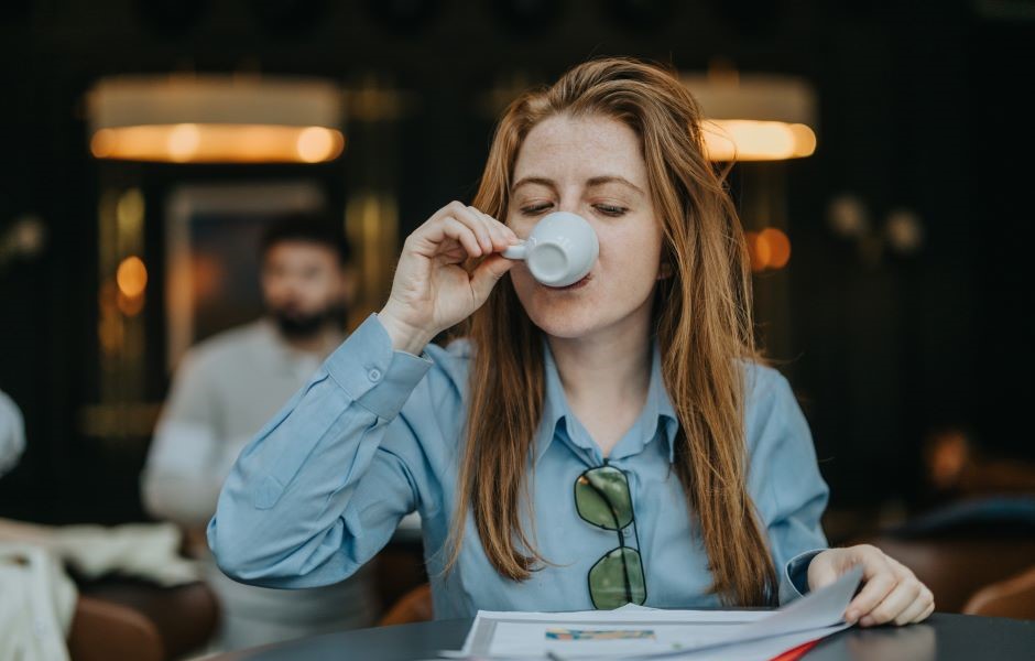 Mulher com cabelo castanho, usando blusa azul e óculos de grau presos à camisa, tomando uma xícara de café ou chá em um ambiente aconchegante, com foco na expressão de satisfação. Ao fundo, pessoas estão em um espaço moderno e bem iluminado, criando uma atmosfera de cafeteria ou sala de reunião, ideal para quem busca conforto e relaxamento.