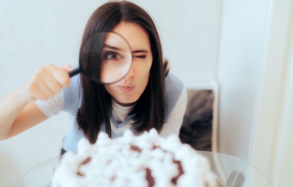 Jovem mulher com cabelo castanho escuro usando uma camiseta cinza clara, observando uma sobremesa com uma lupa, em ambiente claro e minimalista.