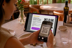Uma cliente sentada à mesa de um restaurante usa o celular para escanear um QR Code exibido em um tablet. A tela do tablet mostra uma mensagem incentivando a avaliação da experiência do estabelecimento no Google.
