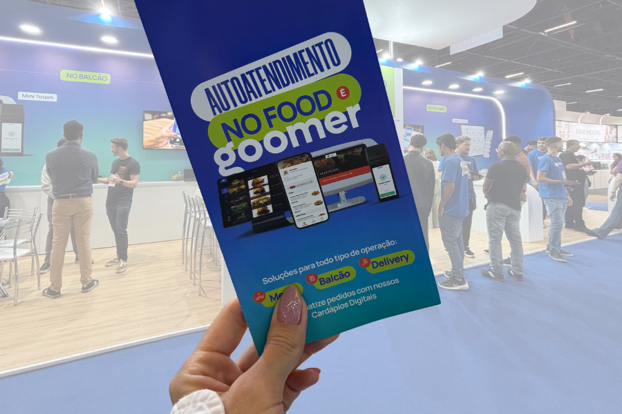 Banner de autoatendimento do NO FOOD e GOOMER em evento de tecnologia, com várias pessoas interagindo com os stands no fundo.
