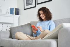 Jovem sorridente usando celular enquanto relaxa no sofá de casa, ambiente moderno e aconchegante, ideal para o conforto e entretenimento.