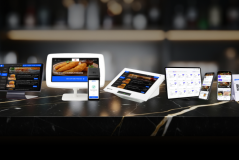 Diversos dispositivos digitais exibindo plataformas de pedidos online de alimentos, incluindo tablets, smartphones e computadores, enquanto estão dispostos em uma mesa de mármore escura em um ambiente moderno.