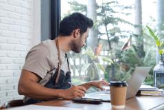 Homem estudando e fazendo anotações com laptop, celular, caneta e café na mesa de um espaço moderno, com vista para a natureza através de uma grande janela.