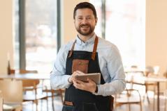 Homem sorridente usando um tablet em um ambiente de café, vestido com avental, mostrando a modernidade e a gestão de estabelecimentos comerciais.