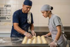 Profissionais de pastelaria/padaria trajando aventais, gorros de proteção e luvas transparentes organizam porções redondas de massa sobre tabuleiro metálico. Na bancada de inox sobressai farinha polvilhada, enquanto ao fundo observa-se parede revestida a azulejos claros com cartaz informativo.